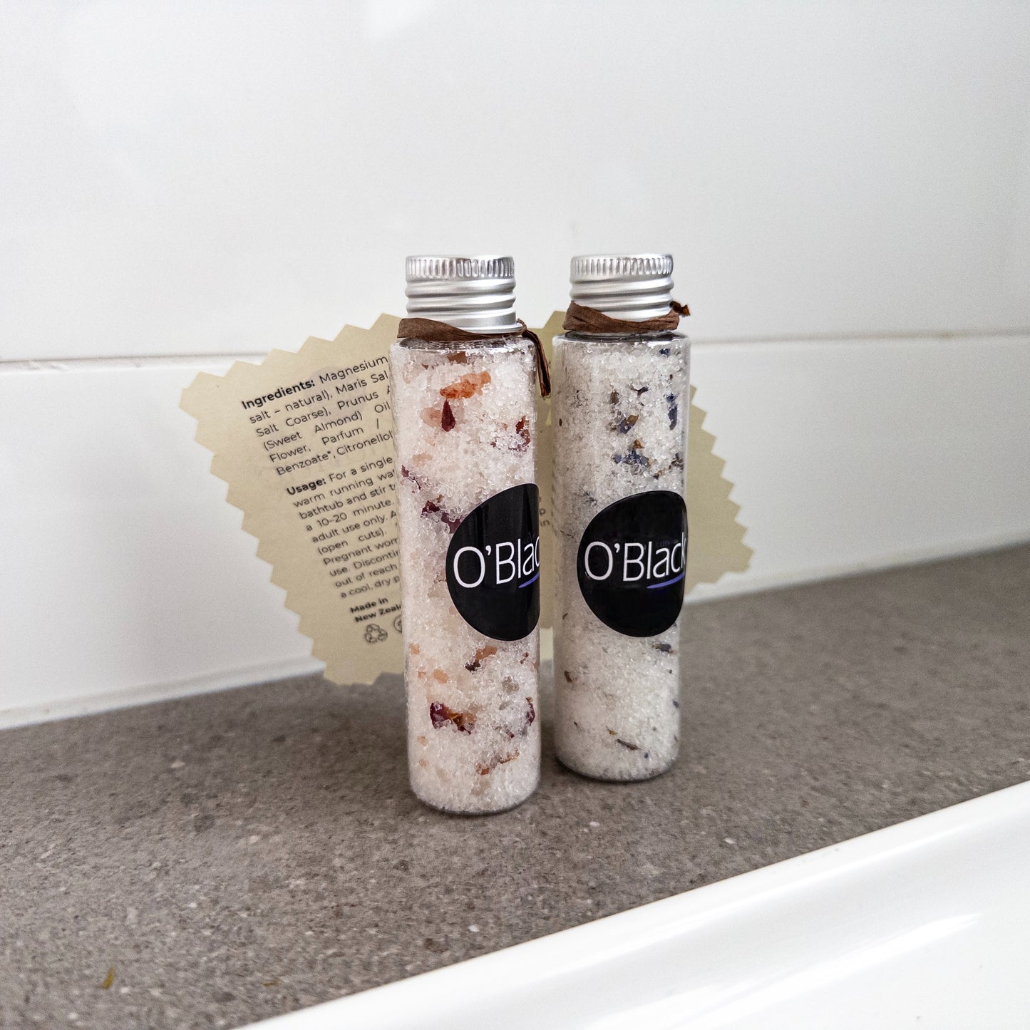 Two bottles of O'Blad bath salts on a bathroom counter with a white tiled wall in the background.