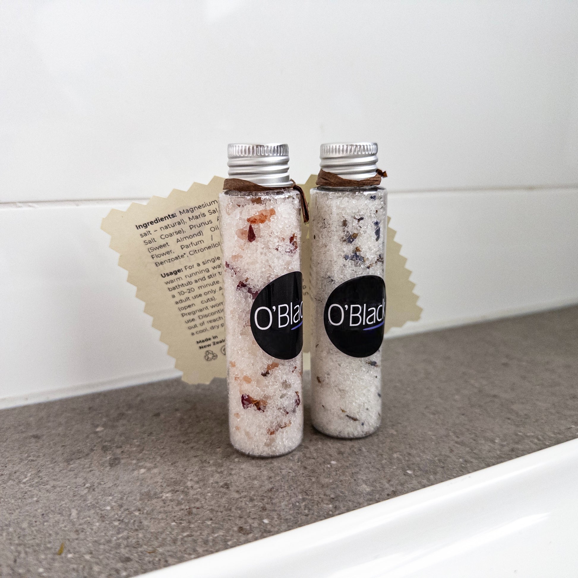 Two bottles of O'Blad bath salts on a bathroom counter with a white tiled wall in the background.